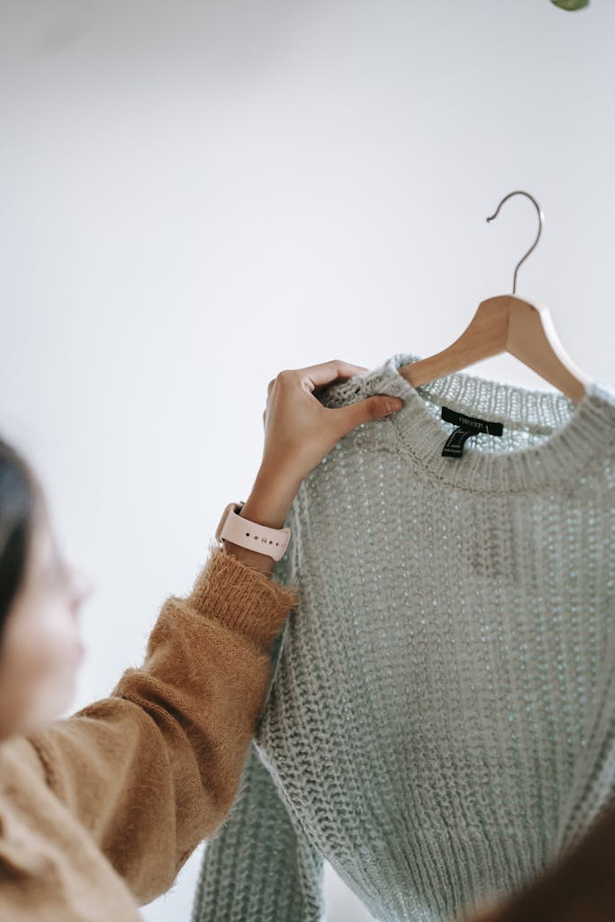 Crop anonymous female designer in casual clothes demonstrating new sweater on hanger while standing in room